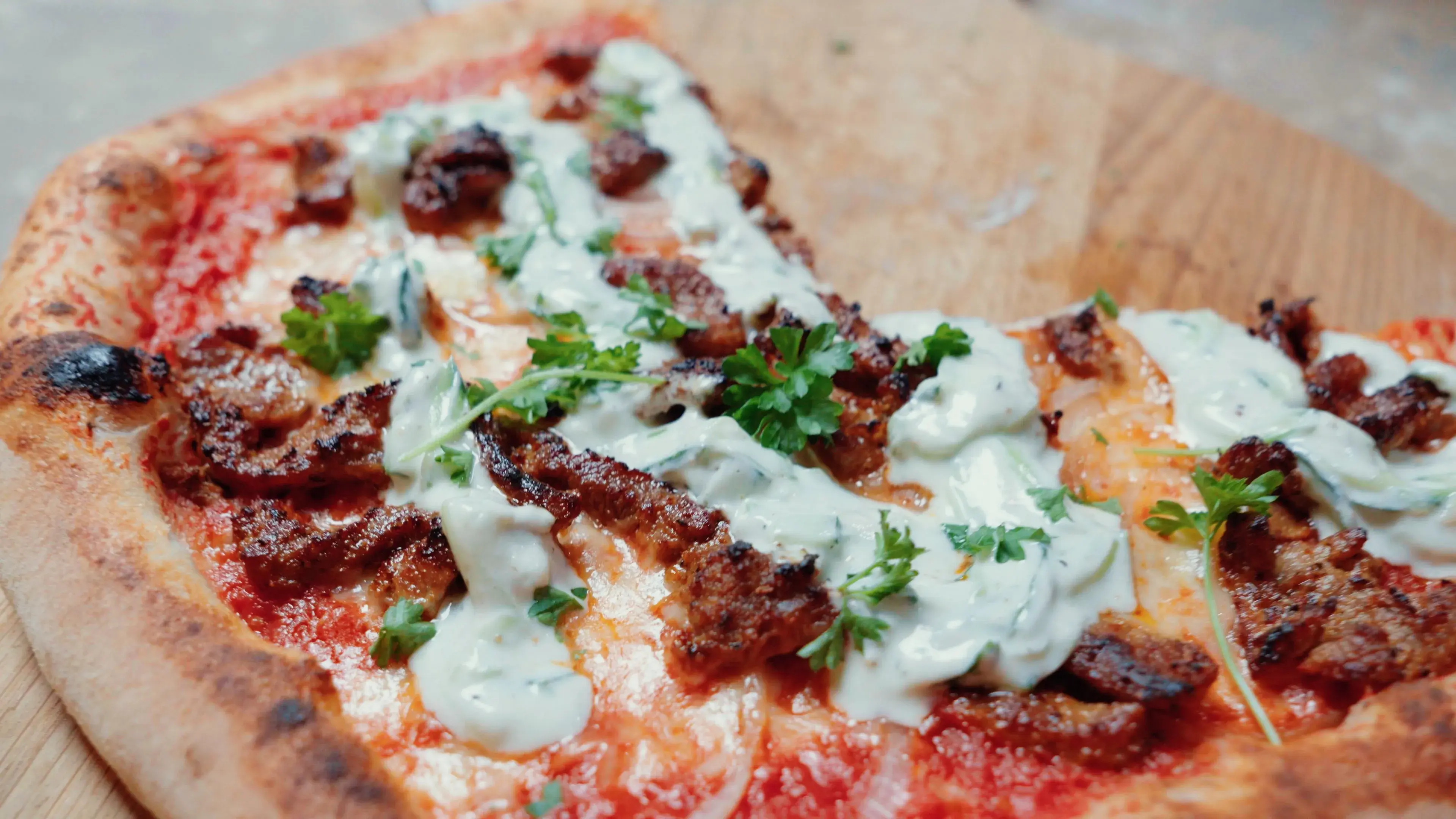 Fertig gebackene Gyros Pizza auf einem Holzbrett mit goldbraunem Rand, Zwiebelstreifen, Pfannengyros und gleichmäßig aufgespritztem Tzatziki sowie frischer Petersilie