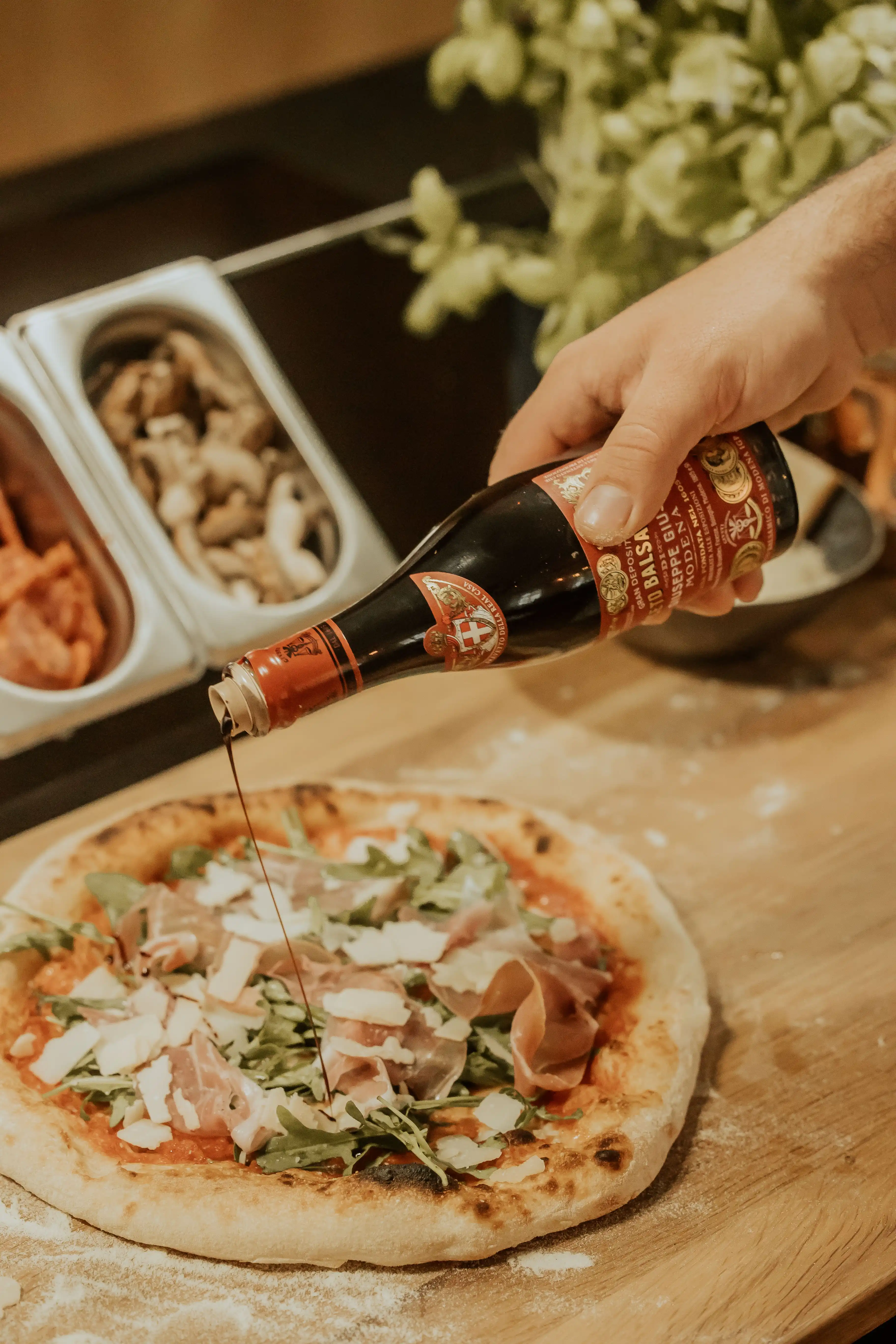 Aceto Balsamico di Modena wird in feinem Strahl über eine Pizza mit Prosciutto, Rucola und gehobeltem Käse geträufelt