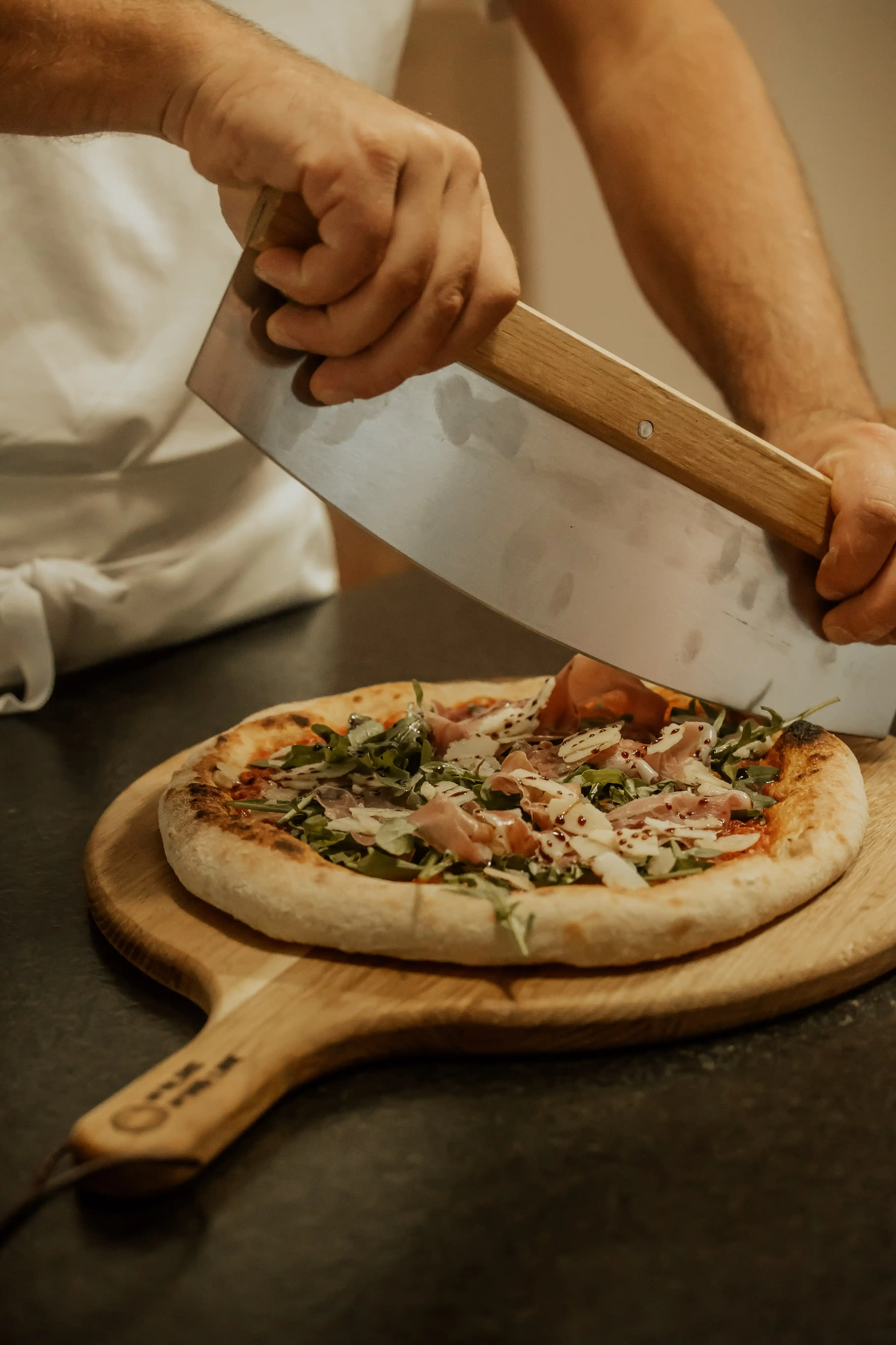 Pizzabäcker belegt eine gebackene Pizza mit Rucola und Parmaschinken vor einer Belegstation mit Tomatensoße und Fior di Latte